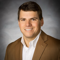 Daniel White, a man with short brown hair, smiles at the camera. He is wearing a light brown blazer over a white collared shirt. The background is a neutral gray.