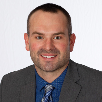 Lance Ibeling, a man with short dark hair and a beard, smiles at the camera. He is wearing a gray suit, a blue shirt, and a blue plaid tie against a plain white background.