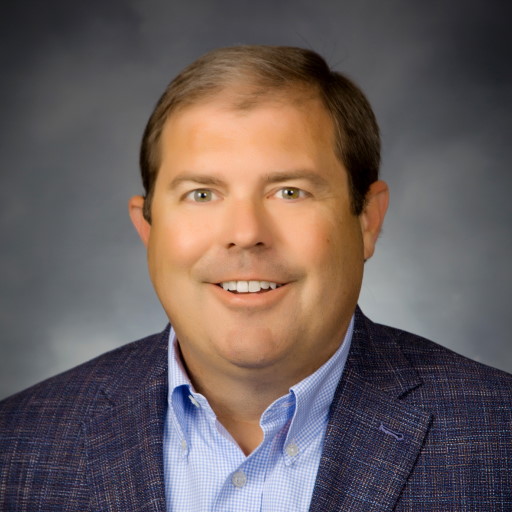 Trent Hurley, in a suit jacket and blue shirt, smiles warmly against a dark, neutral background.