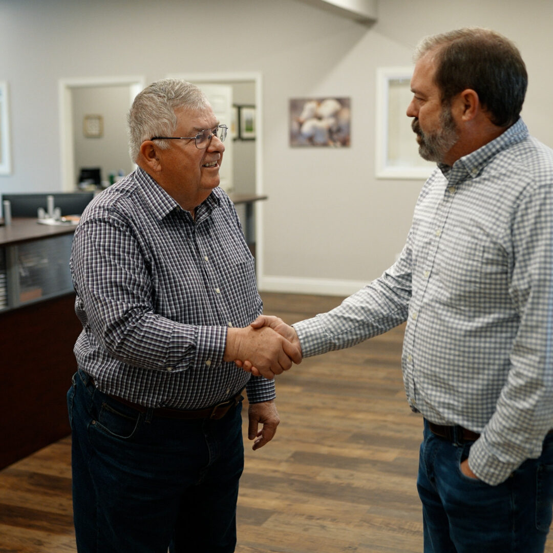Two men in casual shirts shake hands in an office setting. The background shows a reception area with a person working at a desk. The room has wooden flooring and simple décor with framed pictures on the wall.
