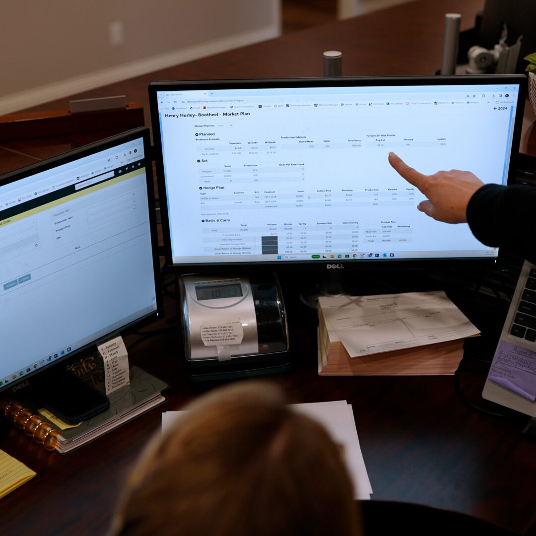 A person points at data on one of three computer monitors arranged on a desk. The screens display spreadsheets and software interfaces. Office supplies and notes are scattered on the desk, and a small plant is visible in the background.