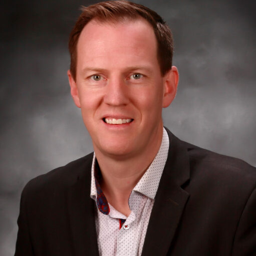 Aaron Erickson, with short hair, stands smiling in a dark suit and light shirt against a gray, cloudy background.