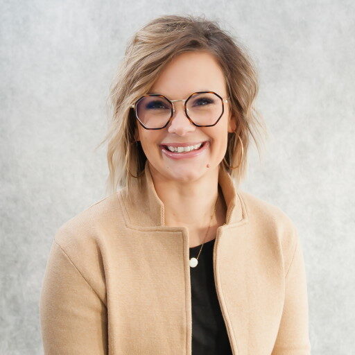 A person with shoulder-length light brown hair and round glasses smiles at the camera. They channel a touch of Annie Oakley-Huber style, wearing a beige jacket over a black top against a gray, neutral background.