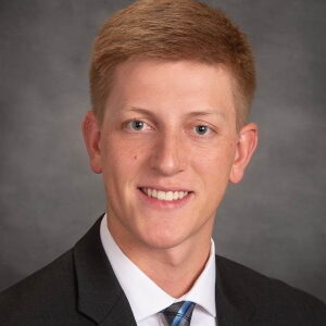 Joseph Prohaska, a person with short light brown hair, is seen wearing a dark suit, white shirt, and striped tie, smiling warmly against a plain gray background.