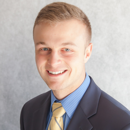 A young man, perhaps Cal or Noah, with short blonde hair is smiling. He wears a dark suit jacket over a blue dress shirt, paired with a yellow striped tie. The backdrop is light gray.