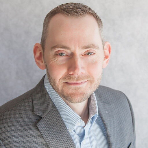 Jared Knudson, a man with short hair and a beard, stands out in his grey suit jacket and light blue shirt, smiling warmly against a light grey background.