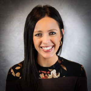 Portrait of Liz Waletich with long dark hair, wearing a black top adorned with floral patterns. She smiles warmly against a neutral gray background.