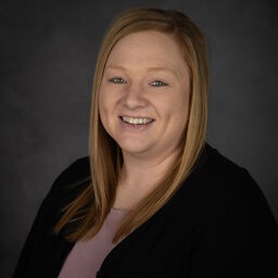 With long, light brown hair, McKenzie Smith smiles warmly at the camera. They are wearing a black top over a pink shirt, set against a plain, dark gray background.