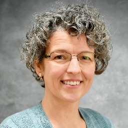 Mindy Whitehill, with her curly gray hair and glasses, smiles warmly in a light blue blazer against a neutral gray background.