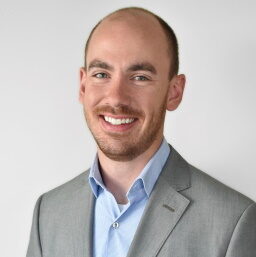 A smiling man, who could be Bobby Reifenrath, with a shaved head and beard, wearing a light blue shirt and gray suit jacket, stands against a plain white background.