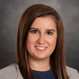 Ellen Schlechter, a woman with long brown hair, smiles against a plain, dark background. She is wearing a gray blazer over a blue top, exuding both confidence and warmth.