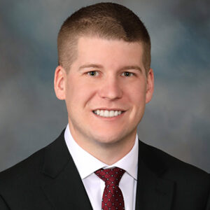 Jackson Waage, in a black suit and red tie, is smiling against a blurred gray background. His short, light brown hair is perfectly styled.