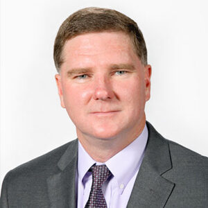 Portrait of John Miller, a man with short brown hair, wearing a gray suit, light purple shirt, and patterned tie. The background is plain white.