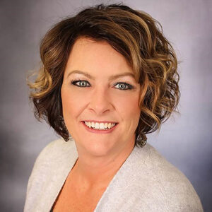 Julie Loebs, with her short, curled hair and light-colored blazer, exudes a warm smile against the neutral background.