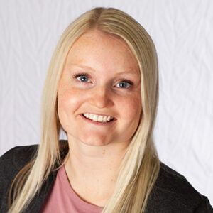 Kayla Smith smiles brightly, showing off her long blond hair as she dons a pink top and dark jacket against the crisp white background.