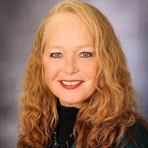 This portrait of Lana Wietgrefe captures her smiling at the camera, showcasing her long, wavy blonde hair. She wears a dark green top and a necklace against a neutral background, exuding an effortless charm.