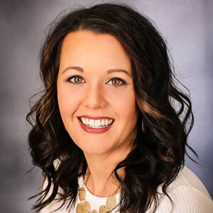Portrait of Laura Peterson with long, dark wavy hair and highlights, wearing a white top and a decorative necklace. She is smiling against a neutral, blue-gray background.