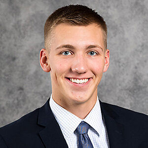 A young man with short hair, identified as Matt Kunerth, smiles broadly. He dons a dark suit jacket, checked shirt, and a patterned tie. The background is a textured gray.