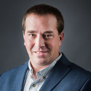 Paul Van Nurden, a man with short brown hair, smiles slightly while wearing a blue blazer over a white checkered shirt. The gray background is softly blurred.